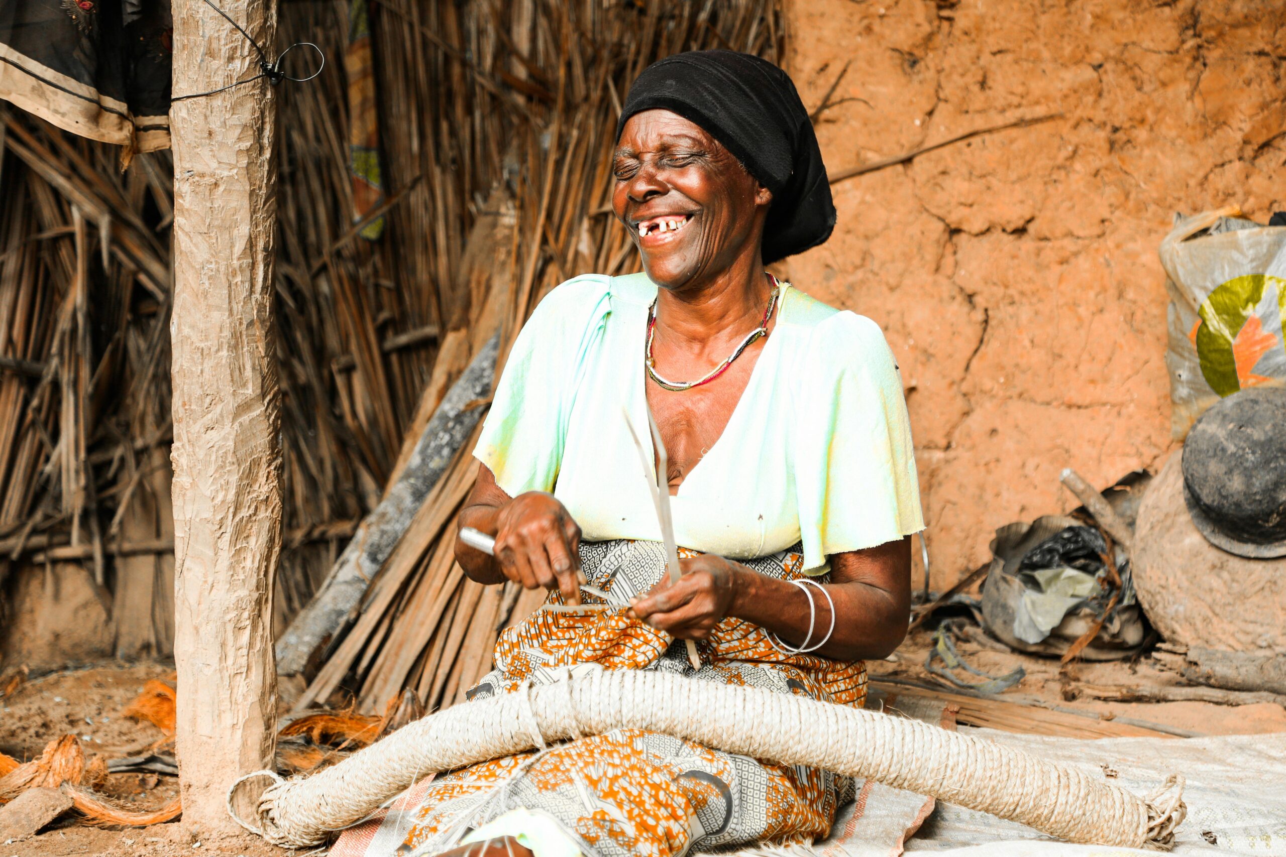 An African woman smiles as she weaves in a rural Tanzanian setting, showcasing traditional craftsmanship.