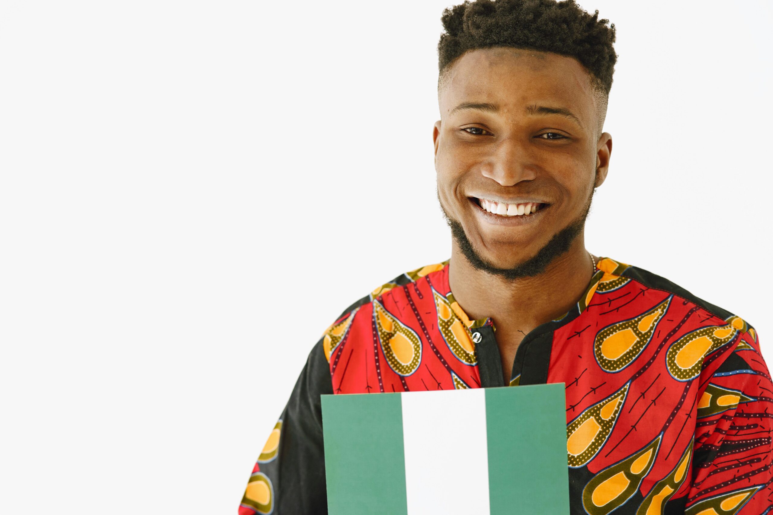 Smiling African man in vibrant shirt holding a Nigerian flag on a white background.